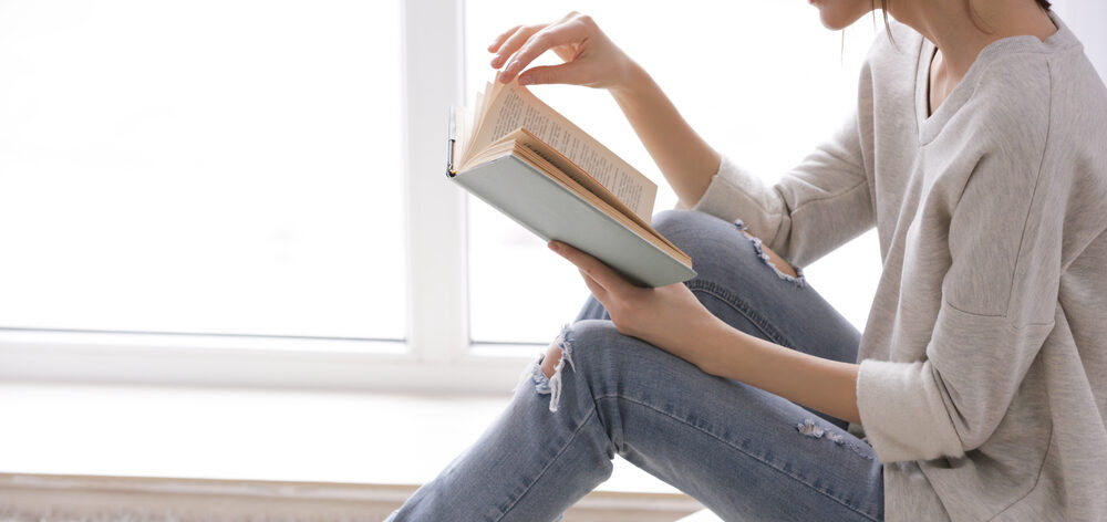 woman turning page in book while sitting by window