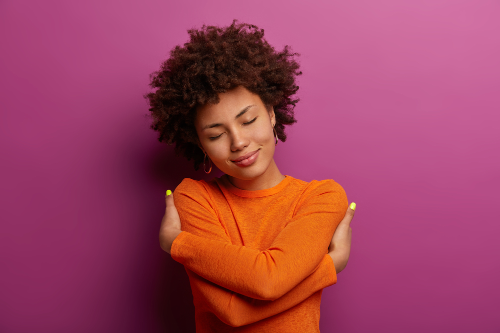 woman in orange shirt hugging herself against a pink background